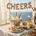 Luxury seaside celebration featuring champagne bottles in an ice bucket, sparkling wine glasses, and a gourmet seafood platter with oysters and shrimp, set against a summer ocean background with "CHEERS" balloons.