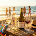 Chilled white and red wine bottles on a sun-drenched beach table during golden hour, overlooking the ocean waves and summer vacationers. The Eastern CEllars